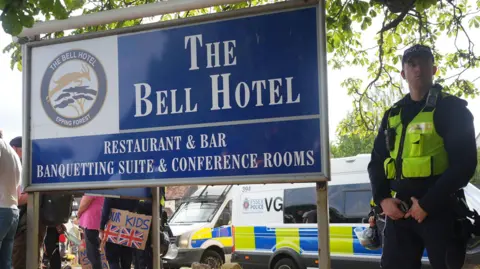 PA Media A police officer standing next to a large blue sign that reads "The Bell Hotel". Two police vans are parked behind him and a crowd of protesters are also gathered.