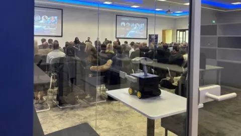 Janine Machin/BBC The picture shows a conference room with dozens of people sitting in chairs looking to the front of the room. Five people are in chairs looking at the audience with two large screens behind them on a white wall. The screens have a partial photograph of the earth from space on them with pictures in circles laid across the background image. The photo of the room is taken from outside, and so a large glass window and door are in the foreground of the image.