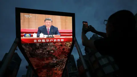 Reuters A person uses her mobile phone near a giant screen showing Chinese President Xi Jinping delivering a speech