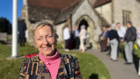 Hannah Gray/BBC Virginia Lewis-Jones looks at the camera smiling. She is standing outside a church where people in the background can be seen standing and talking. The sun is shining.