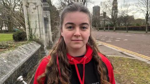 Katie Martin looking at the camera. She is holding what looks like a trumpet or a similar instrument. She has a hoodie on that is branded with the Cardiff Jazz Society logo.