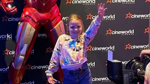 Ellie is stood with one hand almost on her hip and the other hand up in the air, she is smiling and posing for a camera in front of a superhero with a Cineworld banner behind her.