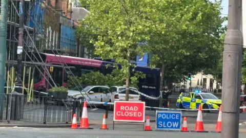 Katy Lewis/BBC A wider view of the crashed bus in the building. A red sign in front of it reads 'Road closed' with another blue sign reading, 'Police slow'. Red cones can be seen blocking the road. A marked police car with its lights flashing is parked to the right of the bus.