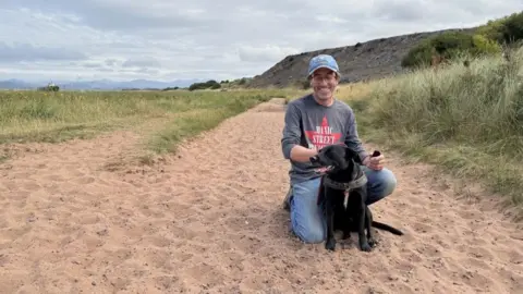 Andrew Tresise kneeling behind his seated dog - a black labrador - on a large sandy path with rough grass to either side, and a bluff behind him with a view of mountains in the distance. He is smiling, wearing glasses, jeans, a blue denim baseball cap and a grey sweatshirt.