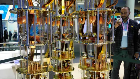Alberto Estevez/EPA/Shutterstock Three golden and copper "chandeliers", festooned with components, hang down vertically. Their bright metal catches light from all directions. A man on the right, wearing a jacket, jeans and open necked shirt, and a conference attendee's lanyard round his neck, is holding up his phone to take a picture of the display. The chandeliers are inside a large conference venue and other people can be seen in the background. 