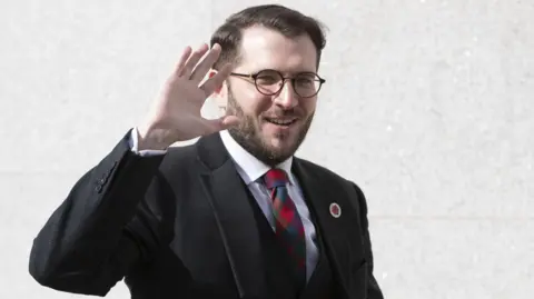 PA Media Paul Sweeney wearing glasses, a dark suit and a patterned tie raises a hand while standing outdoors in front of a light-coloured stone wall.
