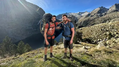 File photo of Aziz Ziriat (left) and Sam Harris in shorts and T-shirts wearing large hiking backpacks and stood on top of a hilltop overlooking a gorge with their arms on each others' shoulders.