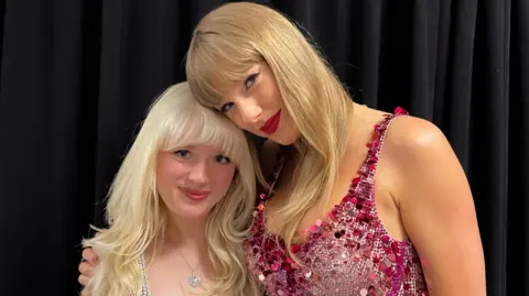 Maisie Peters A short woman with bleached hair in a white sparkly dress stands next to a tall blonde woman in a glittery bodysuit (Taylor Swift)