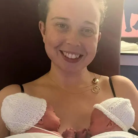 Katie Matthews The picture shows a woman smiling at the camera. She has two new
born babies on her chest. They're wearing knitted hats and are swaddled in a blanket.