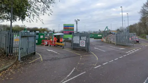 The entrance of the recycling and household waste site which has a silver fence at the perimeter. 