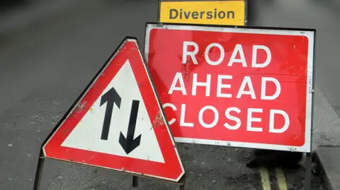A red road closed sign next to a triangular roadworks sign and a yellow diversion sign on a road surface