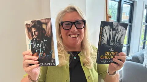 BBC/Lucy Ashton A woman with long blond hair is wearing black rimmed glasses a black top and lime green cardigan. She is smiling and holding up two paperback books