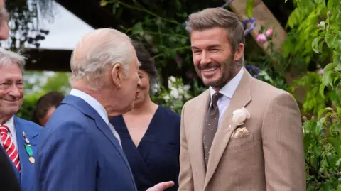 A main with short blonde hair and a light brown suit jacket and brown tie standing in front of a green hedge and talking to a man with short grey hair and a blue jacket