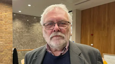 Steve Morphew is wearing a grey/light brown jacket over a navy jumper and red and white checked shirt.