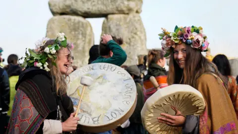 PA Media Two women dressed in ponchos and flower crowns banging on handheld drums at Stonehenge. They are wearing face paint and sunglasses and look very happy.