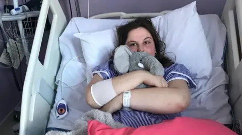 Ella Moulsdale Image of a girl with long brown hair, wearing a blue and white stripe hospital gown who is lying in a hospital bed cuddling a toy elephant.