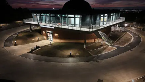 The observatory, seen from outside and from an elevated angle, partly flood lit and partly silhouetted against a sunset