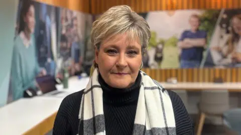 Susan looking at the camera. She has short blonde hair and is wearing a black jumper and a black and white scarf.