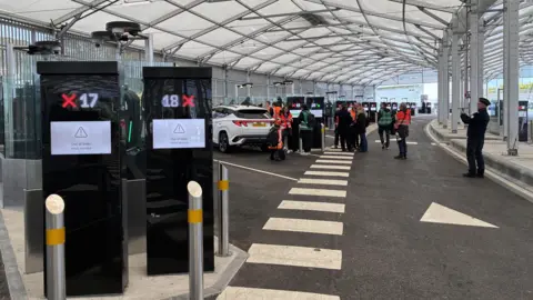 Two finger printing machines in the foreground of a hanger at Eurotunnel in Folkestone with cars and people in the background.