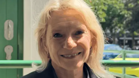 Senator Elaine Millar wearing a dark jacket, standing outside a public toilet entrance, with a green accessible restroom door visible in the background.