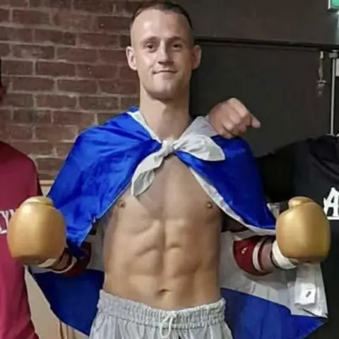 An image of a Sean McInnes, shirtless in white shorts, with a Scotland flag draped around his shoulders and wearing a pair of gold boxing gloves.