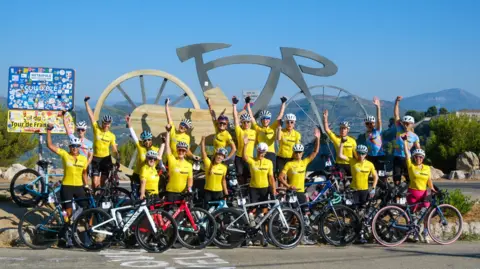 Michelle Sharland The group of amateur cyclists doing the Tour de Femme in France. All the women are wearing yellow t-shirts and black shorts. They each have their arm in the air, and are stood against a giant silver bike statue.