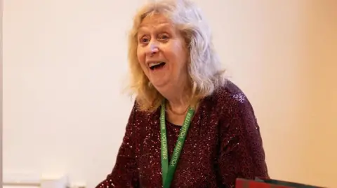 A woman wearing a sparkly dress and green lanyard smiles while looking away from the camera. She is surrounded by gift bags and boxes of toys.
