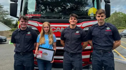 Cambridgeshire Fire and Rescue Three firefighters in blue short-sleeved shirts and trousers, standing with Harriet, who has long, fair hair and is wearing a blue T-shirt and holding her hand-drawn design. All four are standing in front of a bright red and white striped fire engine. 