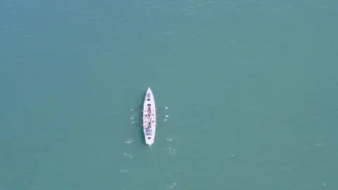 Daniel Stace A rowing boat with four rowers in the middle of the ocean is viewed from the sky.