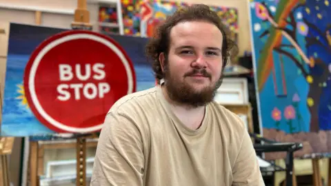 Mackenzie Kane, a young adult, is sitting and looking directly at the camera. Behind him is a wall filled with colourful artwork, including a large hand-painted sign that reads “BUS STOP” in bold letters, reflecting the group’s campaign theme.