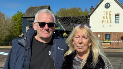 Edd Smith/BBC Mark and Carol Wenn standing in front of a police cordon at The Old Mill pub in Thorpe Marriott. Standing on the left is Mark who is wearing a black T-shirt and blue jacket. He is also wearing a pair of sunglasses over his eyes. Carol, on the right, is wearing a black and white coat, she has blonde hair and a pair of glasses rested on the top of her head.