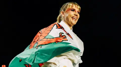 Self Esteem, also known as Rebecca Lucy Taylor, performing on stage at Cardiff Castle. She is wrapped in a Welsh flag, covering her upper body. Her blonde hair is tied up, with her fringe hanging over her forehead. She is smiling and looking away from the camera.
