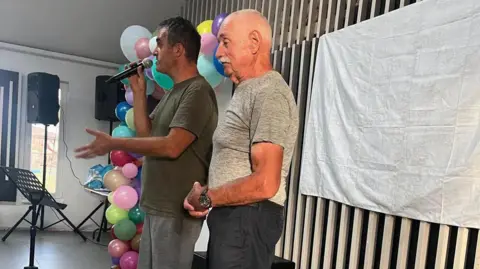 Mustard Seed Jersey Two people stand at the front of an indoor event space, with one speaking into a microphone. Colourful balloons, speakers and a music stand are visible nearby, and a white backdrop hangs against a slatted wall behind them.