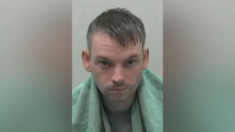 Mugshot of a man with brown hair and wrapped in a green blanket