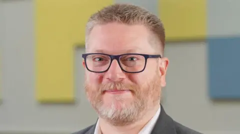 Education Authority A man with short grey hair and a grey beard, wearing a white shirt and grey suit. He is also wearing glasses. 