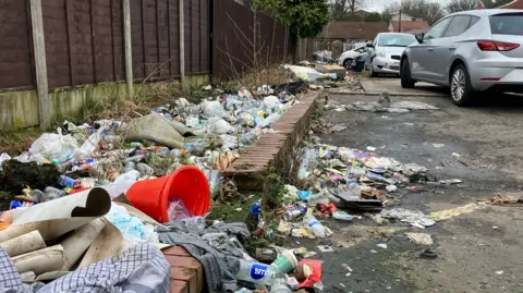 There are piles of rubbish on the ground stretching into the distance. It includes, old rolls of wallpaper, an orange traffic cone, old carpet and scores of bottles and cans. 