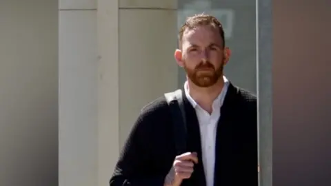 Jamie Niblock/BBC A slightly blurry long shot of Steve Figg at the corner of a building. He has short mid-brown hair and beard and is wearing a black cardigan over a white shirt, with a black bag over his right shoulder.