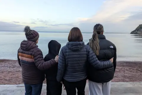 Laura, James, Beth and Kate all stand side by side with their backs to the camera, looking out toward a calm sea from a shoreline.
