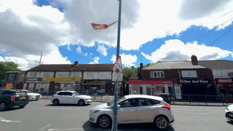 A photo of Princes End which shows several cars passing in both directions. In front of the photographer is a lamppost with a torn St George's flying in the wind. Behind the busy road is a row of retail businesses.