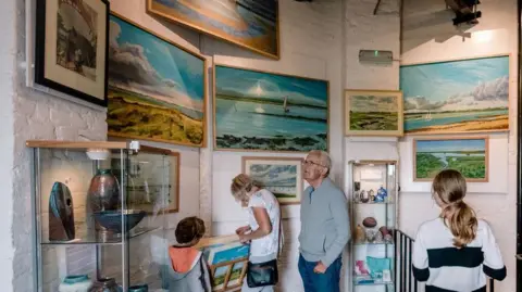 Lucy J Toms Visitors inside the Naze Tower. They are looking at various paintings of the coastal scenery on the wall. There are also glass cabinets with artefacts, including a vase, inside.