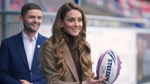 PA Media The princess standing outdoors in what appears to be a sports stadium or training ground. She is holding a white and purple rugby ball branded with the name STEEDEN, positioning it with both hands as if preparing to pass or demonstrate a technique. She is dressed in a brown tweed-style blazer over a high‑neck top, and her long, wavy hair falls over her shoulders.
