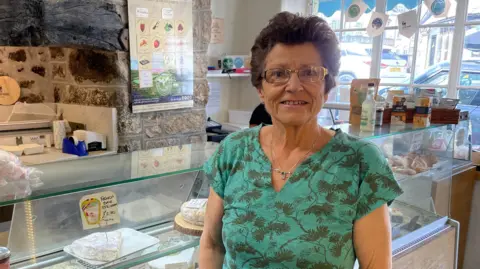 Sue Rogers smiling at the camera. She is stood in front of a counter with food items in it. 