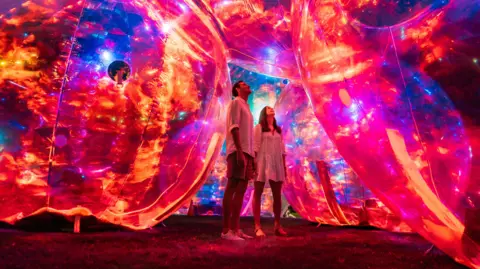 Jesse Lindermann A man and a woman are standing on grass surrounded by giant spheres which are glowing with red, orange, pink and blue lights. They are both looking up and smiling.