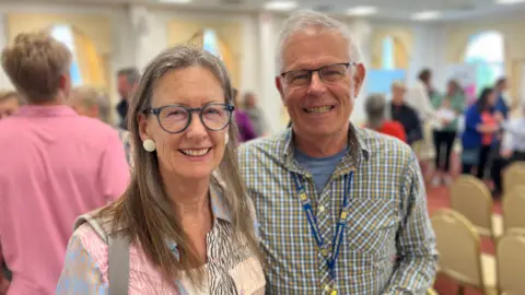 A woman and a man, both wearing collared shirts and glasses, smiling at the camera