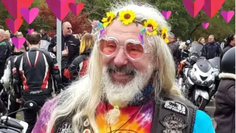 Family handout  Jason Paul Perry pictured with long white hair and a white beard. He is wearing a sunflower headband and heart-shaped glasses. He is wearing a multi-coloured tie-dye T-shirt and a bikers vest on top with different badges printed all over. 