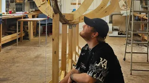 Tooley's Boatyard Jacob Beak, wearing a black t-shirt and cap, is looking up at a wooden boat-like structure. It is in a large workshop.