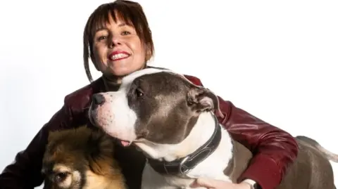 Studio portrait of a person with short dark hair holding two dogs against a plain light background; the person is wearing a red leather jacket and a dark top with a high collar, along with a watch on one wrist. One dog is a large grey-and-white dog with a wide collar, and the other is a smaller brown and black fluffy dog resting in the person’s other arm.