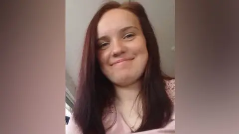 Lucy Campbell A young woman with long, red hair, smiling at the camera, she is wearing a pink top and gold necklace chain.