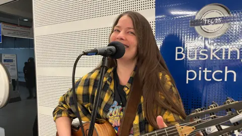 Susana Silva, a woman with long dark hair wearing a black and yellow checked shirt and holding a guitar, smiles as she sings into a microphone in a passageway at Liverpool Street Station. A white wall with with a blue TfL poster saying 'busking pitch' can be seen behind her