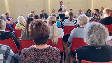 Andy Ralphs standing up to speak in front of the public meeting holding a small diary. He is wearing a white shirt and glasses. The backs of those in attendance at the meeting sitting on orange chairs can be seen in the fore.